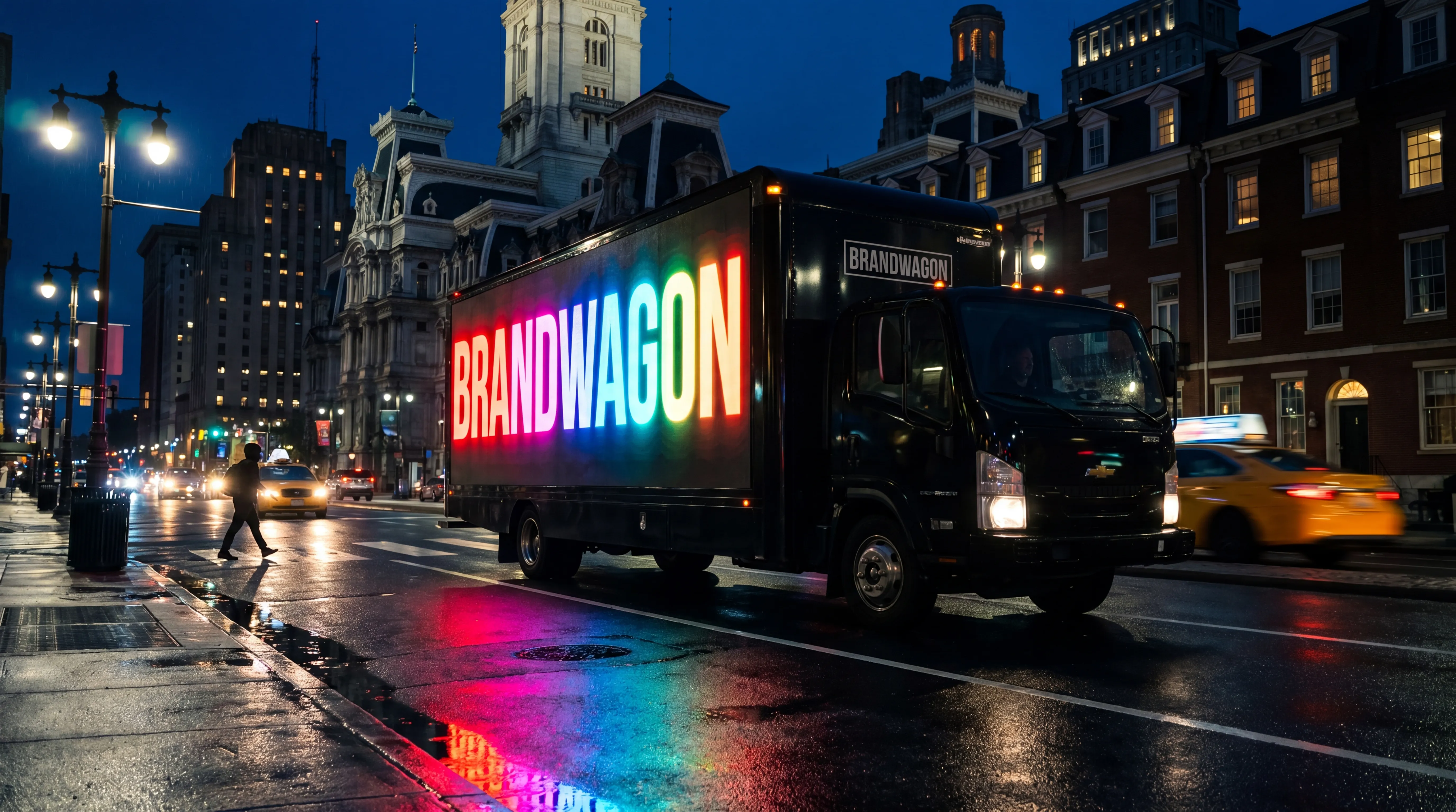 Brandwagon LED billboard truck rolling down Broad Street in Center City Philadelphia at blue hour, rainbow BRANDWAGON wordmark glowing on the screen with neon reflections on wet pavement