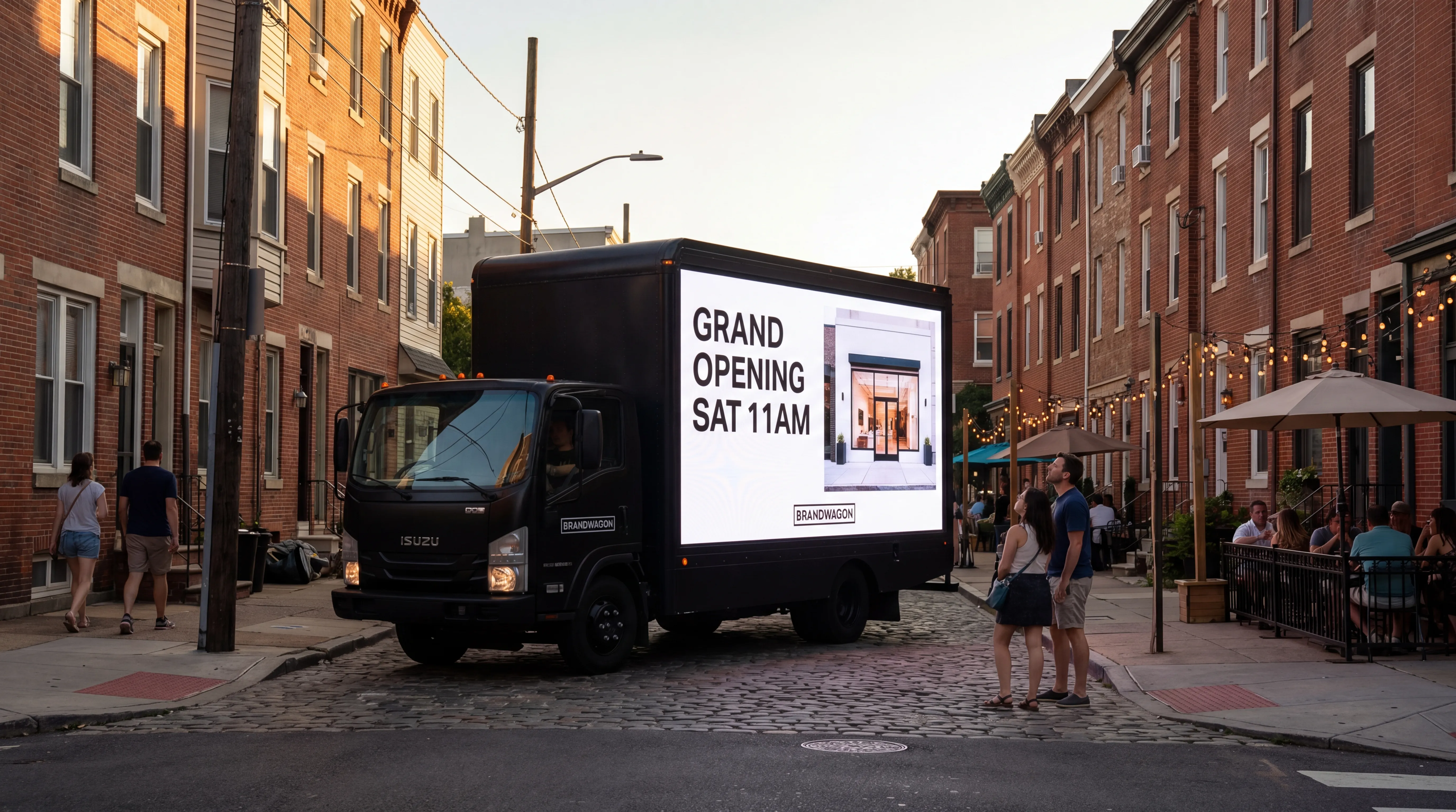 Single Brandwagon LED billboard truck rolling through a Northern Liberties Philadelphia rowhouse street at golden hour, screen showing GRAND OPENING SAT 11AM